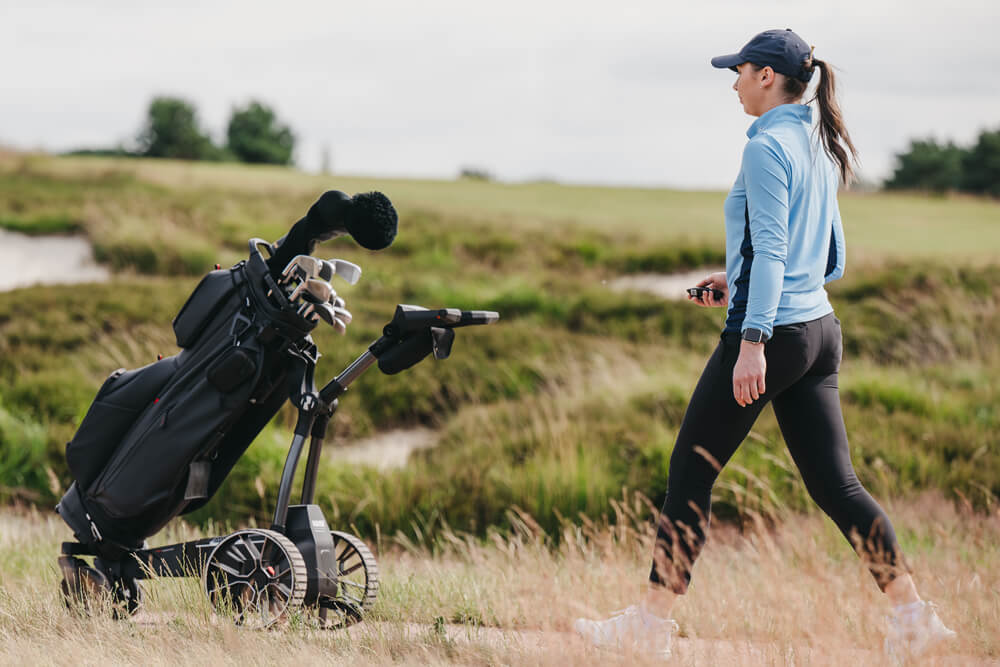 A woman in a light blue long-sleeve top, black leggings, and a navy cap walks beside a black golf bag on the NAVEE Birdie 3 electric golf trolley.